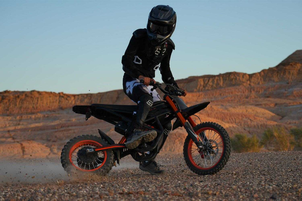 GT54 Electric Mini Motorbike in black and orange with knobby tires, ridden by a person in black protective gear and helmet on rocky terrain