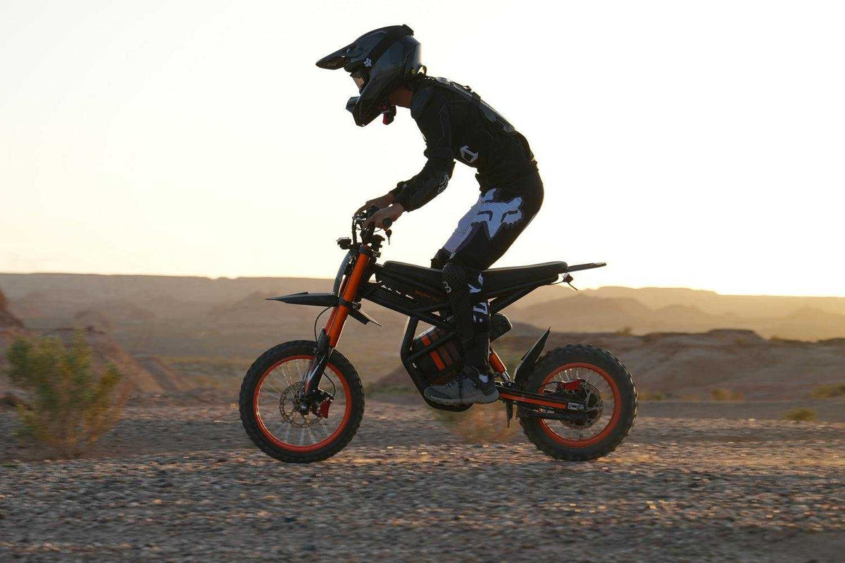 GT54 Electric Mini Motorbike in black and orange with rider in black gear riding off-road terrain at sunset