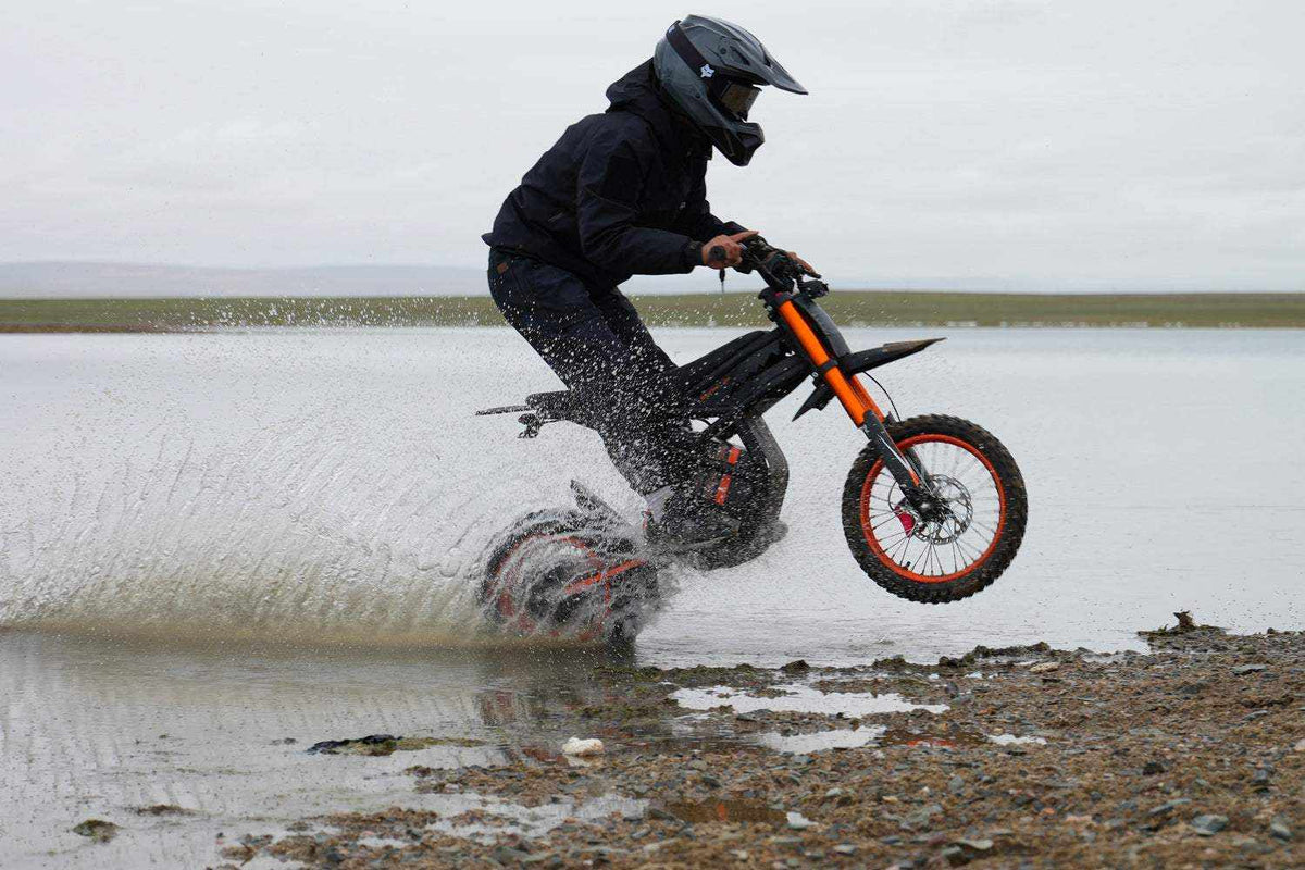 GT54 Electric Mini Motorbike with black and orange frame ridden by person in black gear splashing water on rocky terrain