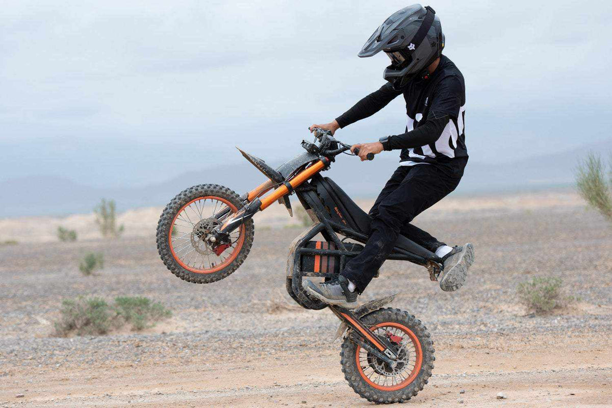 GT54 Electric Mini Motorbike in black and orange with knobby tires performing a wheelie on dirt terrain by a rider in black gear and helmet