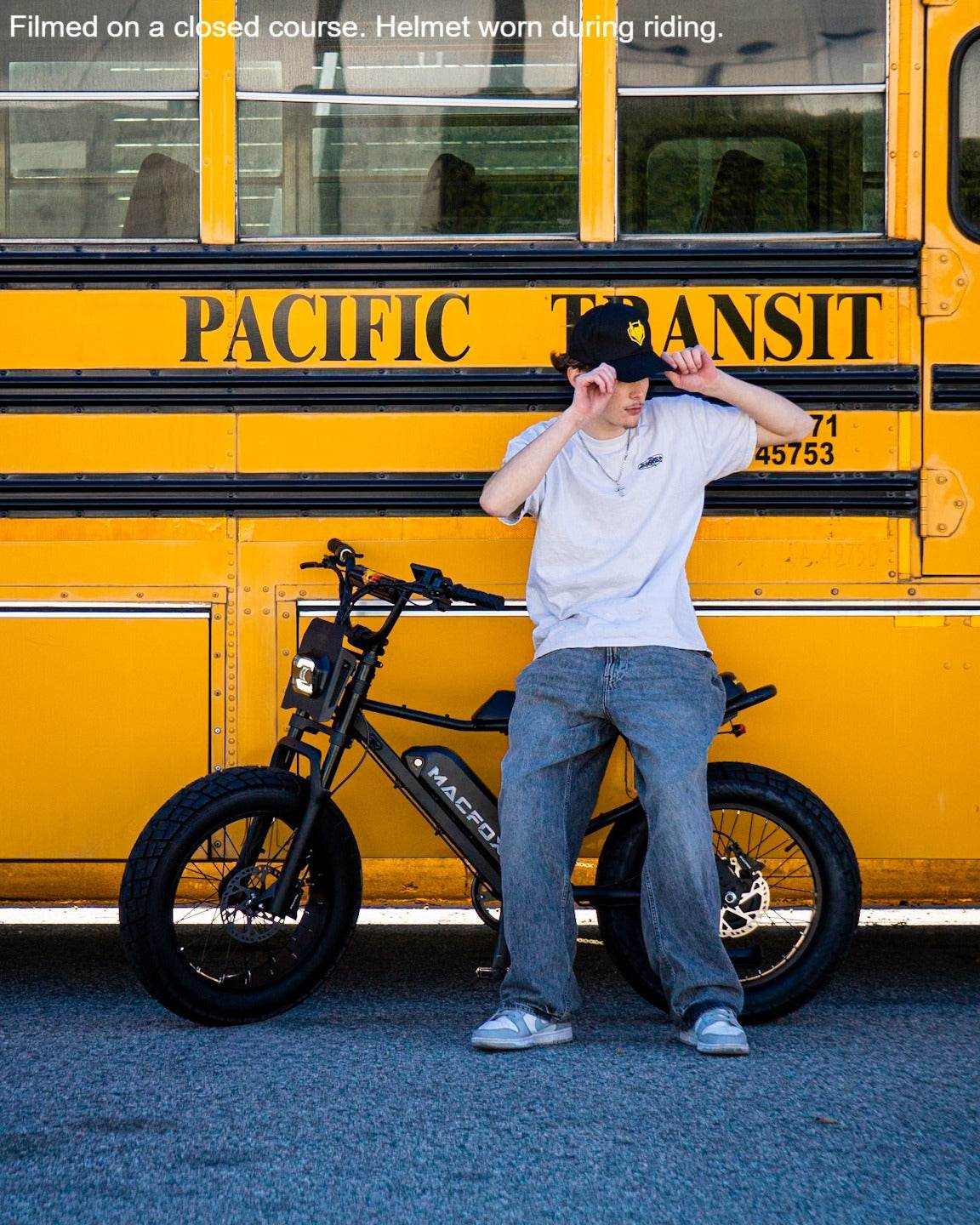 Macfox M16 black electric bike with fat tires, displayed with rider wearing casual grey outfit and baseball cap against yellow bus