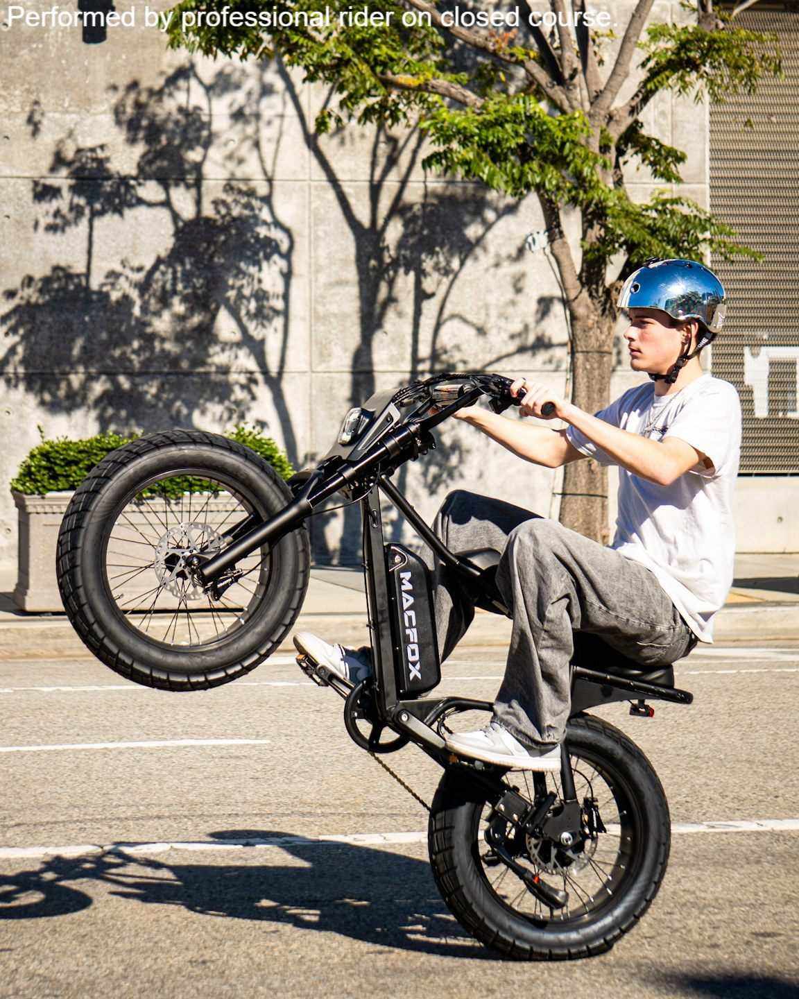 Macfox M16 black electric bike with fat tires and rear hub motor being wheelied by rider wearing blue helmet and casual clothes