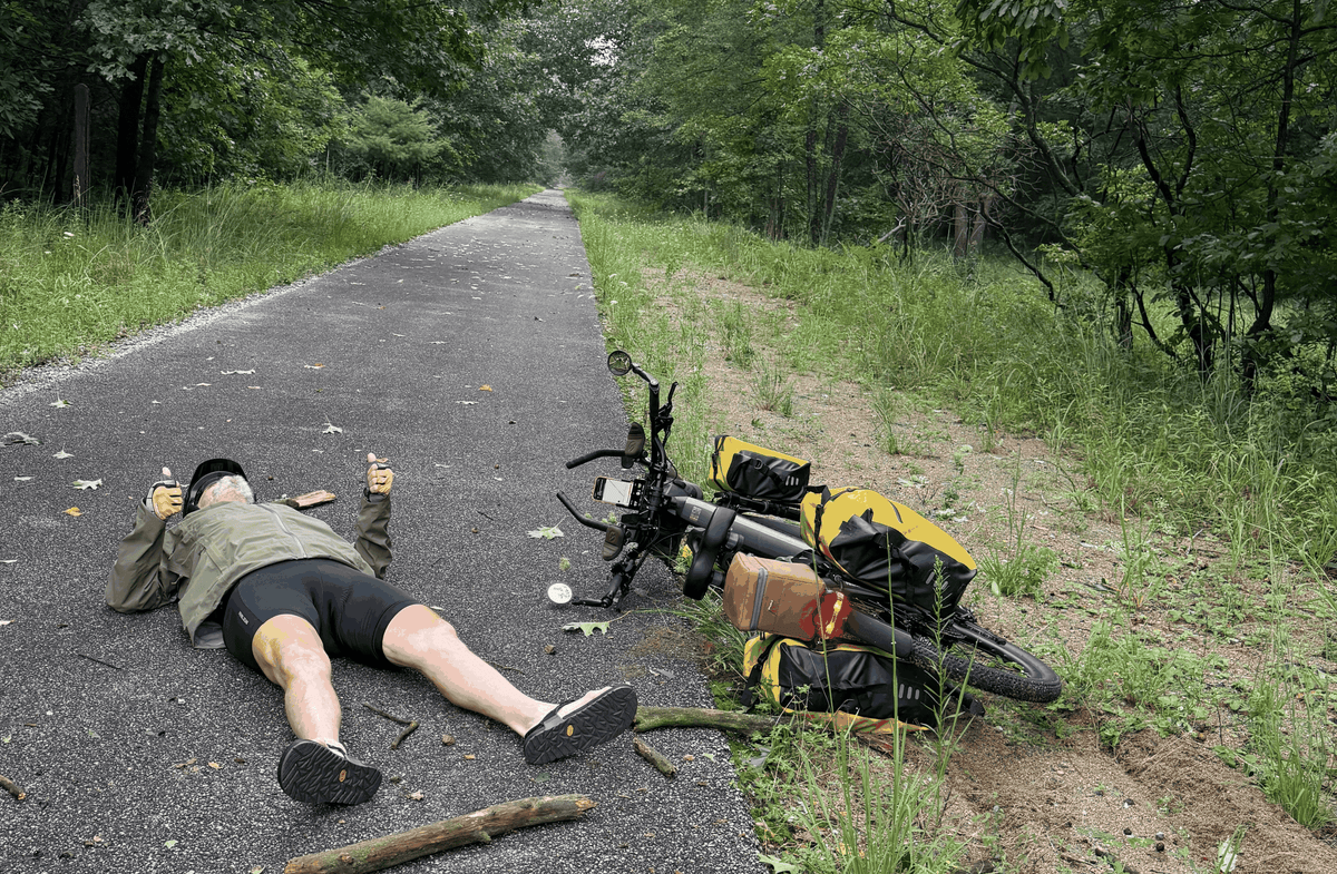 Velotric Discover 3 electric bike in black with yellow panniers and bags, parked on a paved trail next to a person lying on the ground